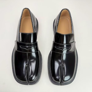 May include: Pair of black leather loafers with a split-toe design. The shoes have a glossy finish, a low heel, and a decorative strap across the top. The interior is a light tan color.