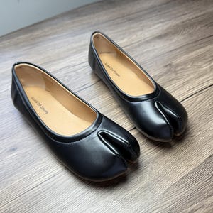 Op de afbeelding: Een paar zwarte leren ballerina's met een split-teenontwerp. De schoenen hebben een ronde neus en een lage hak. De binnenkant is bruin en de merknaam "XUAN DA Shoes" staat binnenin gedrukt.