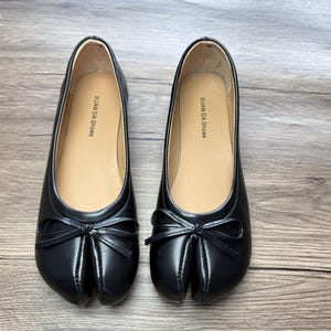 May include: A pair of black split-toe ballet flats with a bow detail. The shoes have a leather-like appearance and a tan insole with the text "XUAN DA Shoes". The shoes are displayed on a wooden surface.