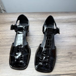 Könnte beinhalten: Paar schwarze Lack-Sandalen aus Kunstleder mit T-Steg-Design und Schnallenverschluss. Die Sandalen haben eine quadratische Zehenpartie und ein strukturiertes Krokodilmuster. Die Absätze sind niedrig und blockartig.