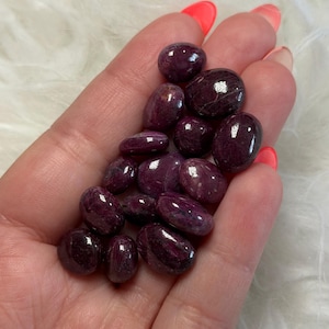 May include: A collection of small, polished, deep magenta and dark purple tumbled stones with smooth, rounded surfaces and subtle natural variations in colour and texture, clustered together to display their glossy, reflective finish.