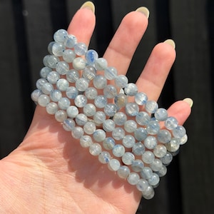 May include: A multi-strand bracelet composed of polished, round, light blue and translucent kyanite beads. The beads feature natural internal striations and varying shades of pale blue, arranged in several parallel rows on an elastic cord.