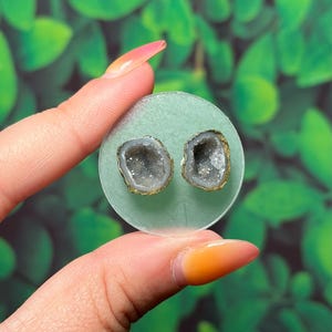 May include: A pair of small, circular stud earrings featuring natural grey geode slices with sparkling crystalline centres. The stones have rough, dark edges and are mounted on a clear, circular backing, showcasing a raw and organic aesthetic.