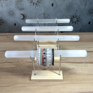 Può includere: Un espositore per gioielli a più livelli con quattro aste orizzontali in selenite bianca sostenute da bracci metallici su basi in legno chiaro. Una collezione di braccialetti di perline in pietre dure è disposta sull'asta anteriore.