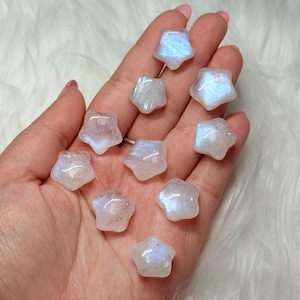 May include: A collection of ten small, star-shaped moonstone carvings. Each piece features a smooth, polished surface with a translucent, milky white appearance and a distinct iridescent blue sheen that shifts when viewed from different angles.
