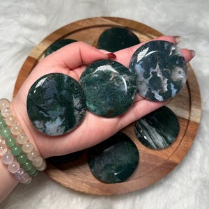 May include: Polished, round moss agate stones in a deep green hue with white and translucent patterns. The stones are displayed on a wooden tray. Each agate stone is approximately 7.6 cm in diameter.