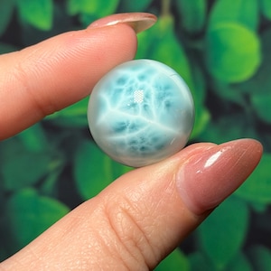 May include: A spherical, polished gemstone, mainly light blue with white veining, held between two fingers. The gemstone has a smooth, reflective surface. The background is a blurred green.