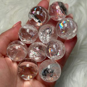 Può includere: Una collezione di sfere di cristallo di quarzo trasparente, ciascuna di circa 2,5 cm di diametro, tenute in una mano. Le sfere mostrano fratture interne e riflessi iridescenti arcobaleno, creando un'affascinante esposizione di luce e colore.