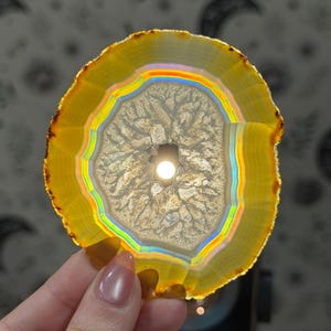 May include: A backlit, circular agate slice with a vibrant yellow and rainbow colour gradient. The stone features a central hole and intricate internal patterns, showcasing natural beauty. The agate is held up against a patterned background.