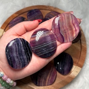 May include: Polished oval fluorite stones in shades of purple, lavender, and pink. The stones are held in a hand, with others on a wooden tray. The stones have a smooth, glossy finish. Ideal for crystal collectors.