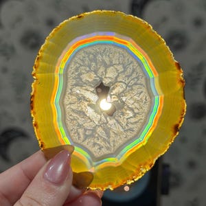 May include: A circular agate slice with a yellow border and a central cavity. The stone has a rainbow-coloured band and detailed internal patterns. The slice is held by a hand.