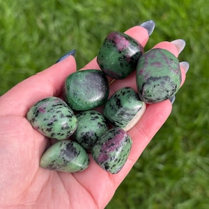 May include: A selection of polished ruby in zoisite stones, each showing a blend of deep green and speckled black with vivid pink inclusions. The stones are smooth and rounded, held in a hand against a backdrop of green grass.