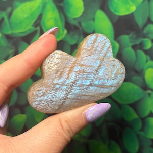 May include: A cloud-shaped gemstone in shades of brown and blue. The polished stone is held in a hand against a background of green leaves. The gemstone has an iridescent quality, with a shimmering appearance.