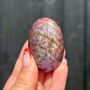 May include: An oval-shaped polished gemstone featuring a mottled mix of deep magenta, soft pink, and translucent white hues. The surface is marked by a network of dark black mineral inclusions and thin, intersecting golden-brown veins throughout.