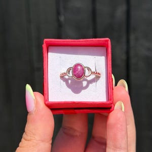 May include: A silver ring featuring a central oval-shaped magenta gemstone. The band has a beaded texture on the sides and is presented inside a small, square, bright red gift box with a white interior lining that holds the ring securely.