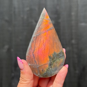 May include: A teardrop-shaped labradorite stone featuring a vibrant orange and pink iridescent flash across its surface. The stone displays natural grey and dark inclusions with a polished finish, showcasing a distinct colour play that shifts in the light.
