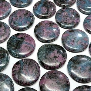 May include: A collection of polished, circular stones featuring a mottled pattern of deep magenta, slate blue, and black mineral inclusions. Each stone has a smooth, reflective surface and a flattened, coin-like shape arranged in a grid pattern.