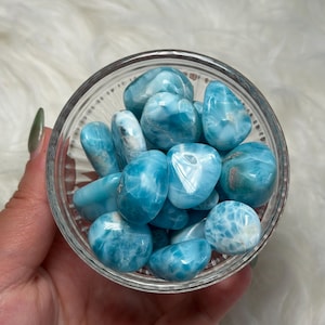May include: A clear glass bowl filled with polished, tumbled larimar stones. The stones are a vibrant blue with white marbling. The bowl is held in a hand, against a soft, white background. The stones have a sea-like appearance.