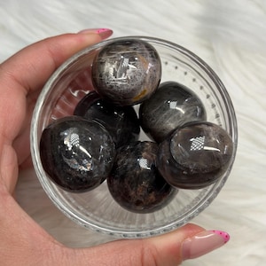 May include: A collection of five polished, rounded stones with a dark, semi-translucent appearance and subtle internal inclusions. The stones are contained within a small, clear, ribbed glass bowl, showcasing their smooth, reflective surfaces.
