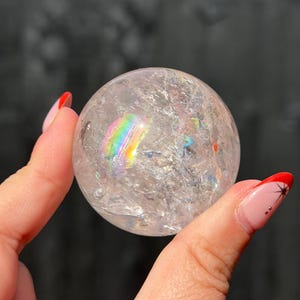 May include: A clear, spherical quartz crystal ball featuring internal fractures and inclusions that create a vibrant, multi-coloured rainbow refraction effect within the stone. The surface of the sphere is smooth and polished, reflecting light.