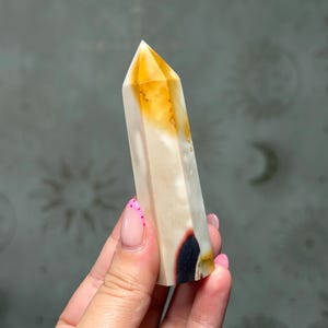 May include: A polished crystal tower with a hexagonal shape, featuring a translucent amber-coloured tip that transitions into an opaque cream-coloured body, accented by a distinct dark brown to black inclusion near the base of the stone.
