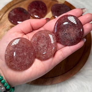 May include: Several polished, reddish-brown strawberry quartz stones are held in a hand. The stones are smooth and rounded, with a translucent appearance. More stones are on a wooden tray. A green and grey beaded bracelet is visible on the wrist.