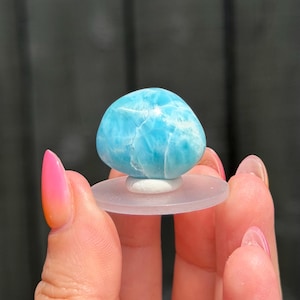 May include: A polished, rounded Larimar gemstone featuring a vibrant sky-blue colour with intricate white, web-like patterns throughout its surface. The stone is mounted on a small, circular, translucent white plastic display stand for stability.