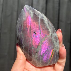 May include: A polished labradorite stone with a teardrop shape, featuring a prominent iridescent magenta and purple flash across its grey surface, accented by dark mineral inclusions and a thin streak of orange near the bottom edge.