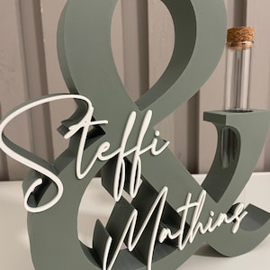 May include: A sage green wooden ampersand sign featuring the white script text Steffi & Mathing attached to the front. The structure includes a cylindrical glass test tube with a cork stopper inserted into the right side of the ampersand.