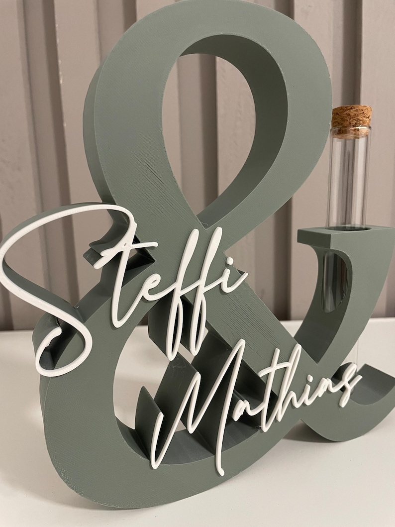 May include: A sage green wooden ampersand sign featuring the white script text Steffi & Mathing attached to the front. The structure includes a cylindrical glass test tube with a cork stopper inserted into the right side of the ampersand.