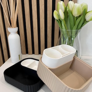 Könnte beinhalten: Zwei dekorative Aufbewahrungsbehälter, einer schwarz und einer beige, mit geriffelten Seiten. Ein weißer Behälter mit zwei kreisförmigen Fächern sitzt auf dem beigefarbenen Behälter. Eine weiße Vase mit Schilf und eine Vase mit weißen Tulpen befinden sich im Hintergrund.