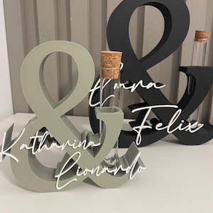 May include: Two ampersand-shaped stands, one sage green and one black, each featuring a clear glass test tube with a cork stopper. The sage green stand displays the white script names Katharina and Lionardo, while the black stand features the names Tanja and Felix.