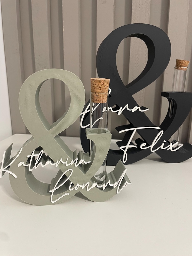 May include: Two ampersand-shaped stands, one sage green and one black, each featuring a clear glass test tube with a cork stopper. The sage green stand displays the white script names Katharina and Lionardo, while the black stand features the names Tanja and Felix.