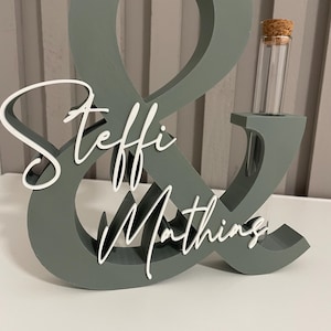 May include: A sage green ampersand-shaped decorative stand featuring the white script text Steffi & Mathing. The structure includes a cylindrical glass test tube with a cork stopper inserted into the right side of the ampersand symbol.