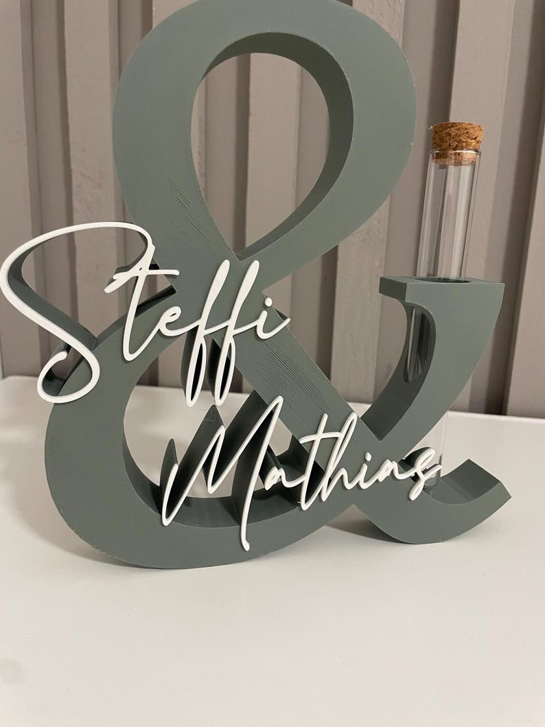May include: A sage green ampersand-shaped decorative stand featuring the white script text Steffi & Mathing. The structure includes a cylindrical glass test tube with a cork stopper inserted into the right side of the ampersand symbol.