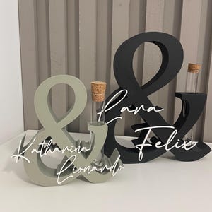 May include: Two ampersand-shaped stands, one sage green and one black, each featuring a clear glass test tube with a cork stopper. The sage stand displays the white script names Katharina and Lionardo, while the black stand displays Lara and Felix.