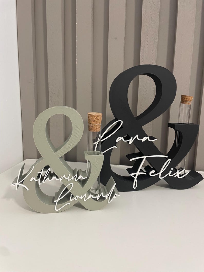 May include: Two ampersand-shaped stands, one sage green and one black, each featuring a clear glass test tube with a cork stopper. The sage stand displays the white script names Katharina and Lionardo, while the black stand displays Lara and Felix.