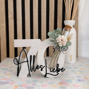 Könnte beinhalten: Eine weiß glitzernde Zahl 70 Dekoration mit dem schwarzen, in Schreibschrift gehaltenen Text Alles Liebe auf der Vorderseite. Ein Glasreagenzglas mit Korkstopfen steckt in der Null und enthält einen kleinen Strauß aus Trockenblumen und Eukalyptus.