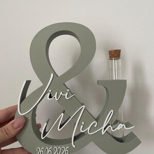May include: A sage green ampersand sign featuring white script lettering that reads Vivi and Micha, with the date 26.06.2026 printed at the bottom. A clear glass test tube with a cork stopper is inserted into the right side of the structure.