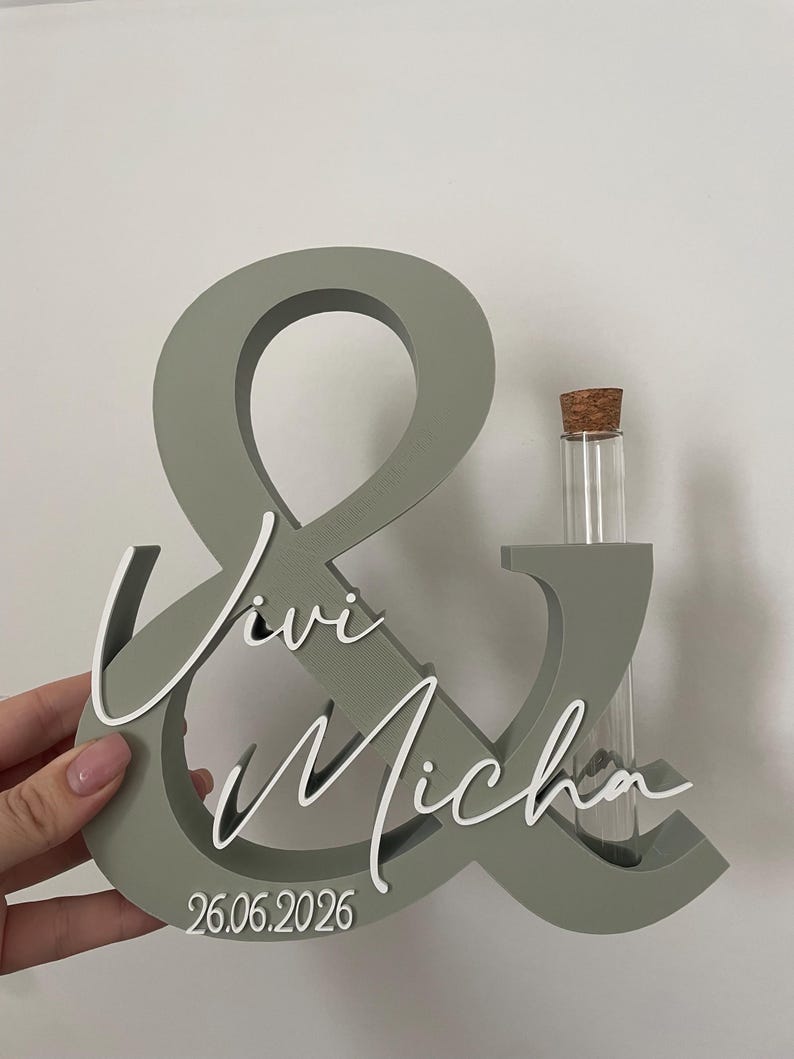 May include: A sage green ampersand sign featuring white script lettering that reads Vivi and Micha, with the date 26.06.2026 printed at the bottom. A clear glass test tube with a cork stopper is inserted into the right side of the structure.