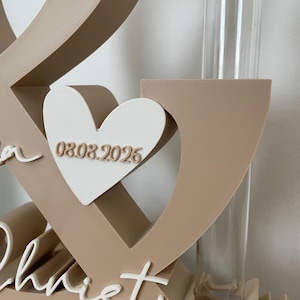 May include: A beige ampersand-shaped display stand featuring a white heart with the date 08.08.2026 in beige lettering. The structure includes a clear glass test tube with a cork stopper and the white cursive name Christian at the base.