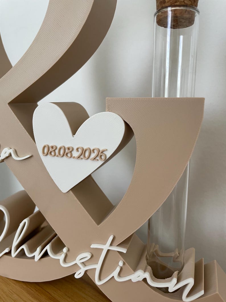 May include: A beige ampersand-shaped display stand featuring a white heart with the date 08.08.2026 in beige lettering. The structure includes a clear glass test tube with a cork stopper and the white cursive name Christian at the base.