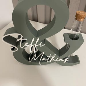 May include: A sage green ampersand-shaped wooden sign featuring the white script names Steffi and Mathing attached to the front. A clear glass test tube with a cork stopper is inserted into a circular cutout on the right side of the ampersand.