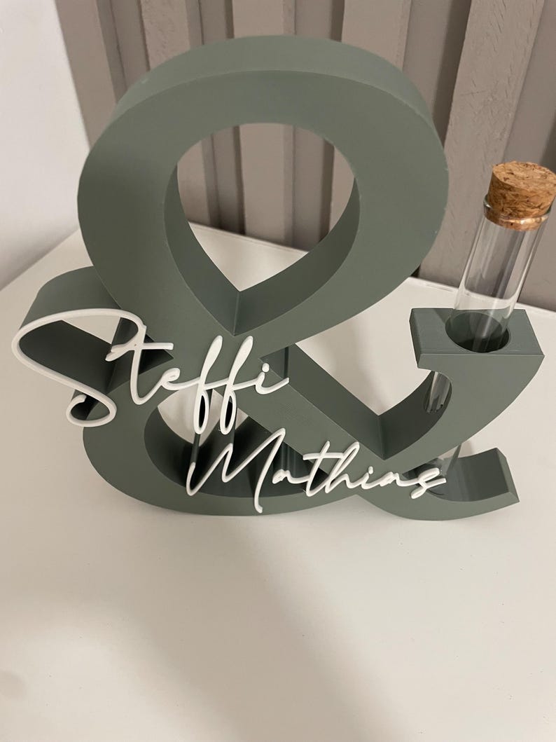 May include: A sage green ampersand-shaped wooden sign featuring the white script names Steffi and Mathing attached to the front. A clear glass test tube with a cork stopper is inserted into a circular cutout on the right side of the ampersand.