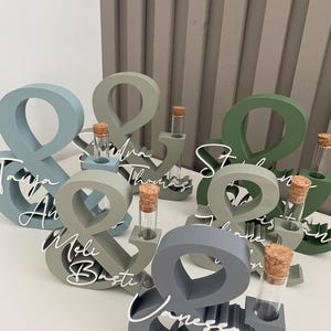 May include: A collection of ampersand-shaped wooden stands in various shades of blue, grey, and green. Each stand features a clear glass test tube with a cork stopper and white script text displaying names such as Tanja, Basti, and Vanessa.