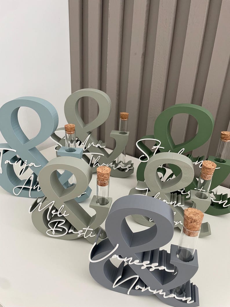 May include: A collection of ampersand-shaped wooden stands in various shades of blue, grey, and green. Each stand features a clear glass test tube with a cork stopper and white script text displaying names such as Tanja, Basti, and Vanessa.