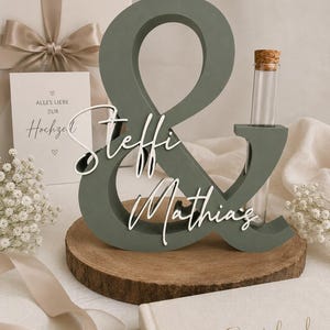 May include: A sage green wooden ampersand sign featuring the white script names Steffi and Mathias attached to the front. A clear glass test tube with a cork stopper is inserted into the top right of the ampersand, which rests on a round wood slice.