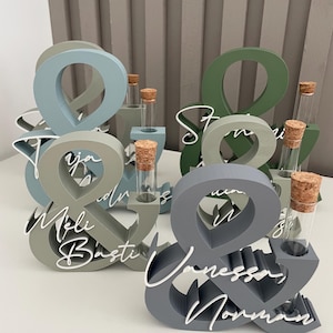 May include: A collection of ampersand-shaped wooden stands in various muted colours including blue, green, and grey. Each stand features a clear glass test tube with a cork stopper and white script text displaying names like Meli, Basti, and Vanessa.