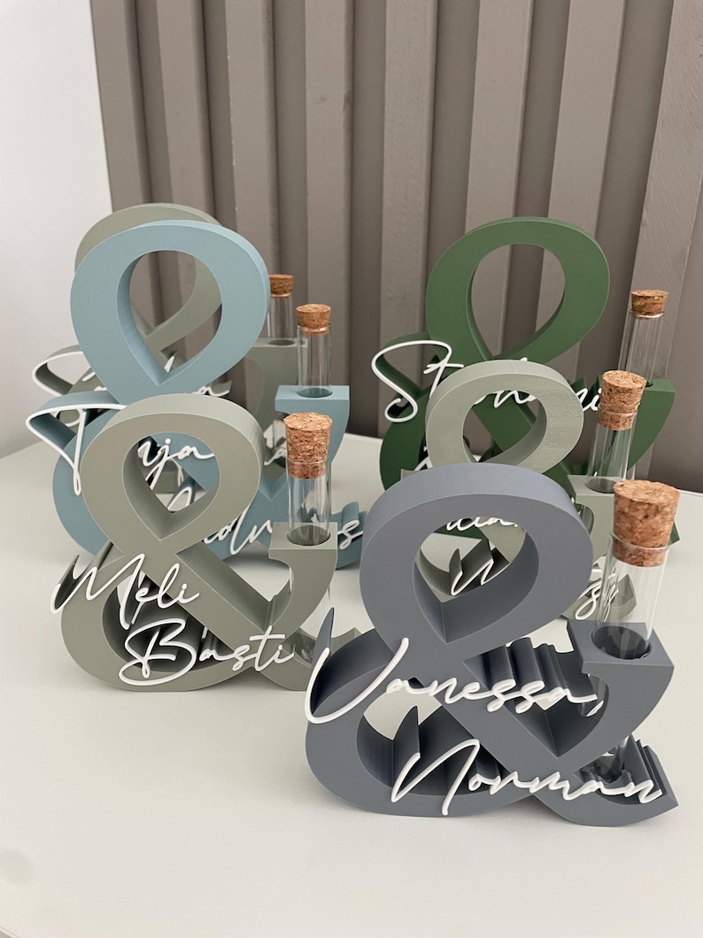 May include: A collection of ampersand-shaped wooden stands in various muted colours including blue, green, and grey. Each stand features a clear glass test tube with a cork stopper and white script text displaying names like Meli, Basti, and Vanessa.
