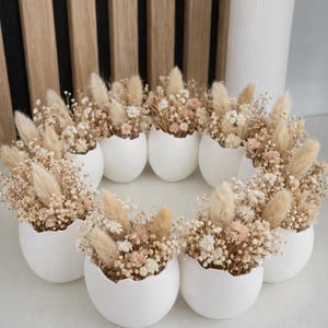 Könnte beinhalten: Eine kreisförmige Anordnung weißer, eiförmiger Keramikvasen, jede gefüllt mit Trockenblumen in Beige, Creme und blassem Rosa. Im Hintergrund steht eine hohe, weiße, gerippte Vase. Die Anordnung befindet sich vor einem hellen Hintergrund.